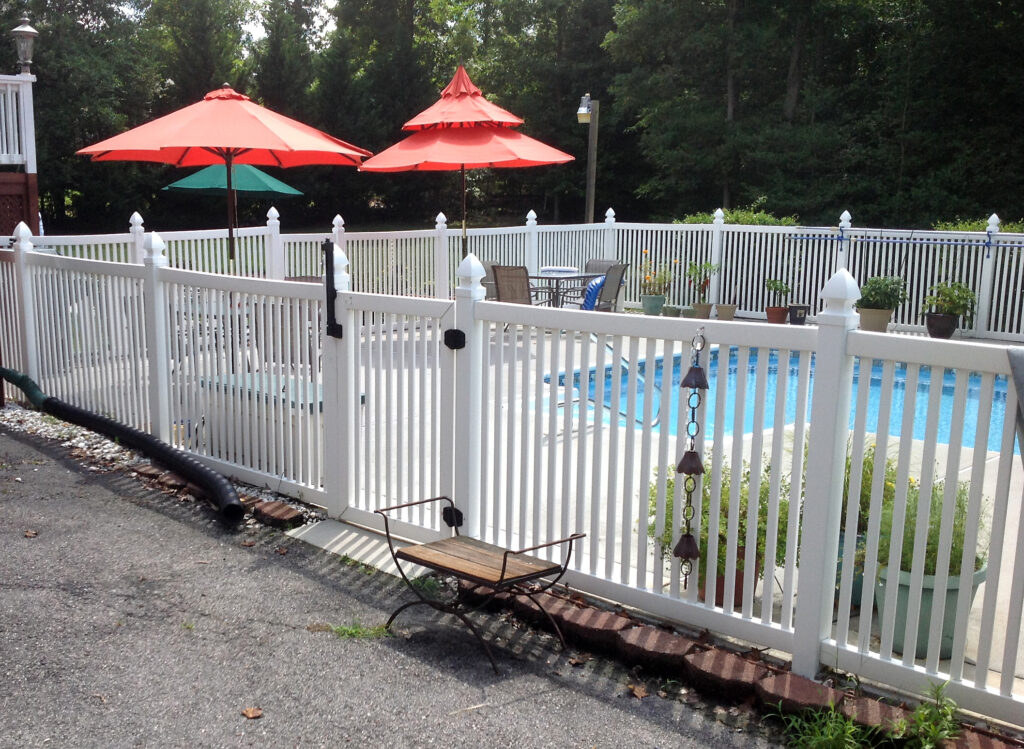 A white picket vinyl fence around a homes backyard pool. The pool fence has large french gothic post caps.